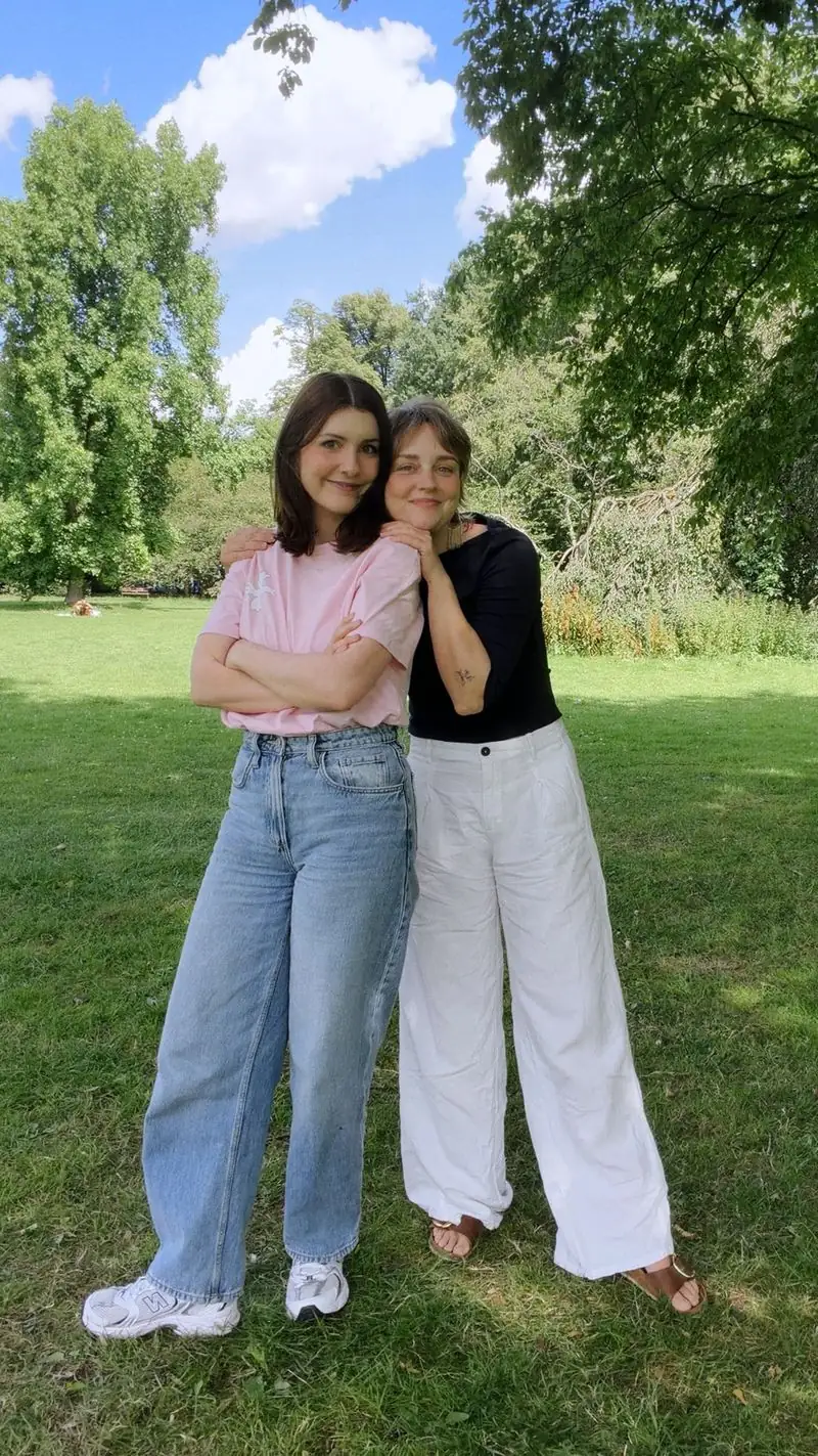 Cristina und Laura stehen Arm in Arm auf einer Wiese im Park