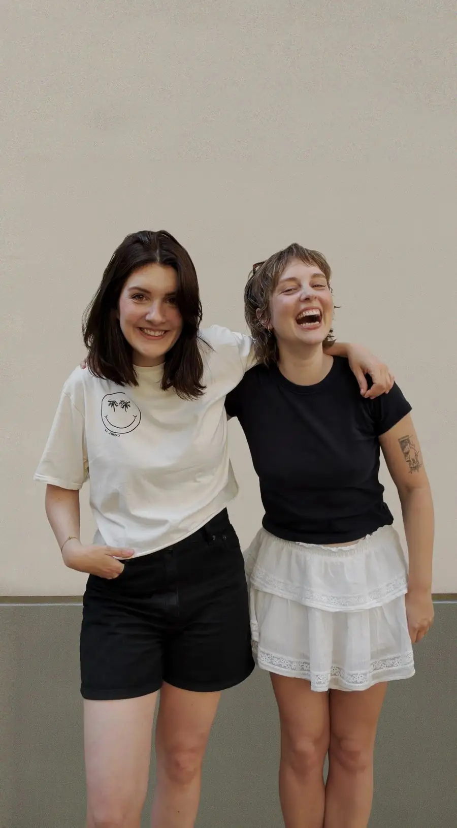 Cristina und Laura lachen Arm in Arm vor einer hellen Wand